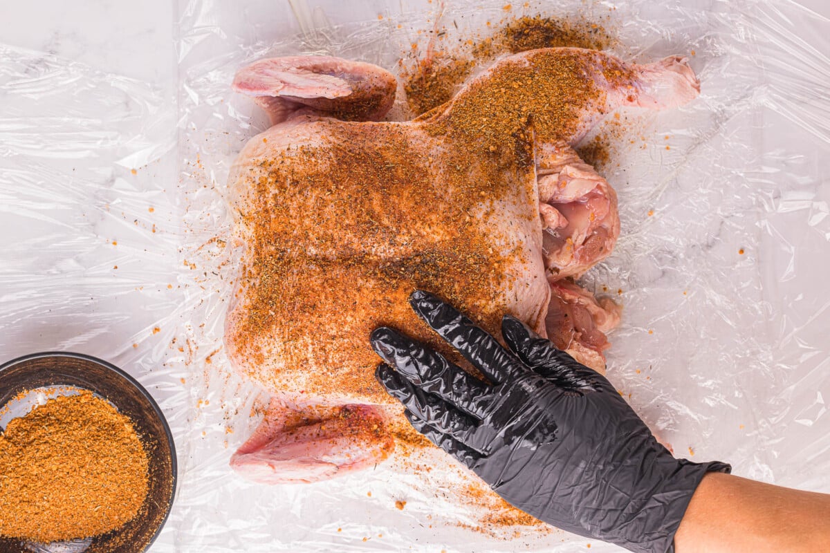 A hand sprinkling the spice blend over raw spatchcock chicken on a baking sheet.