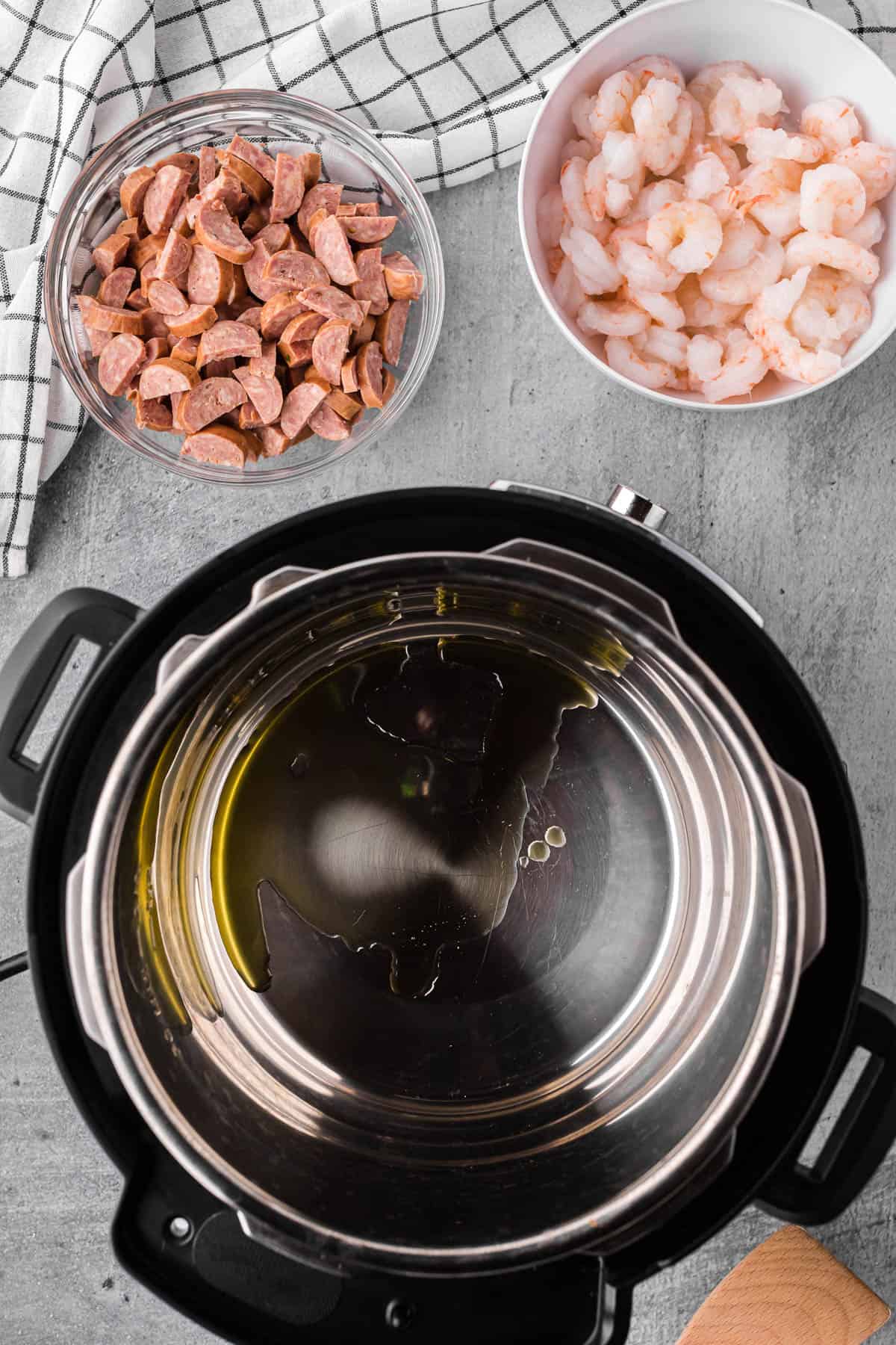 Adding oil to instant pot with bowls of sausage and shrimp for jambalaya recipe.