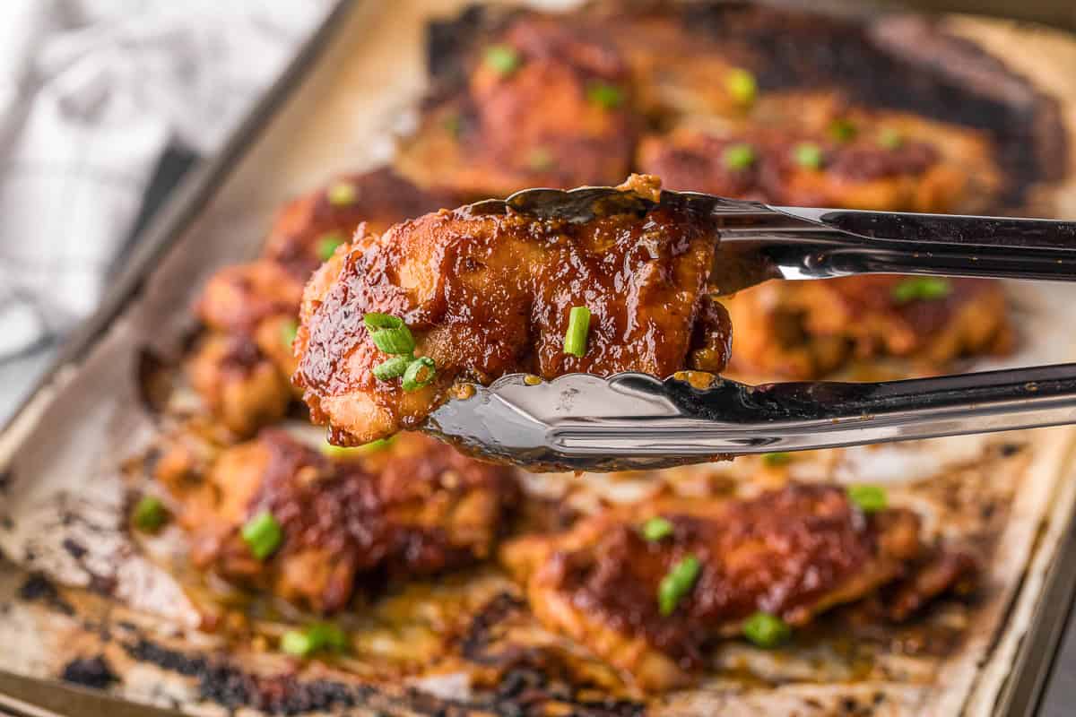 Picking up a apricot glazed chicken thigh with tongs from a baking sheet.