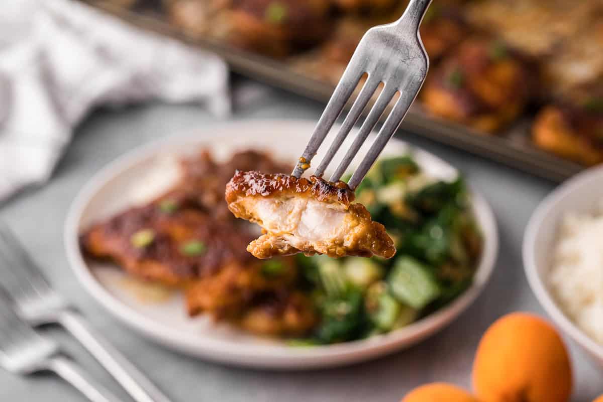 A bite of apricot glazed chicken on a fork with a plated meal in the background.