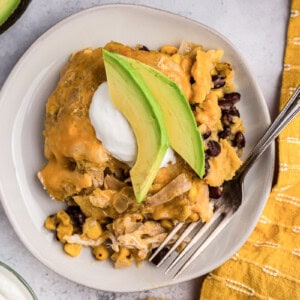 Green Chile chicken enchilada casserole with black beans and corn topped with avocado and sour cream on plate with fork.