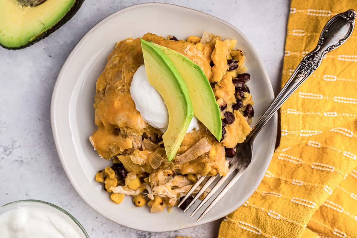 Green Chile chicken enchilada casserole with black beans and corn topped with avocado and sour cream on plate with fork.