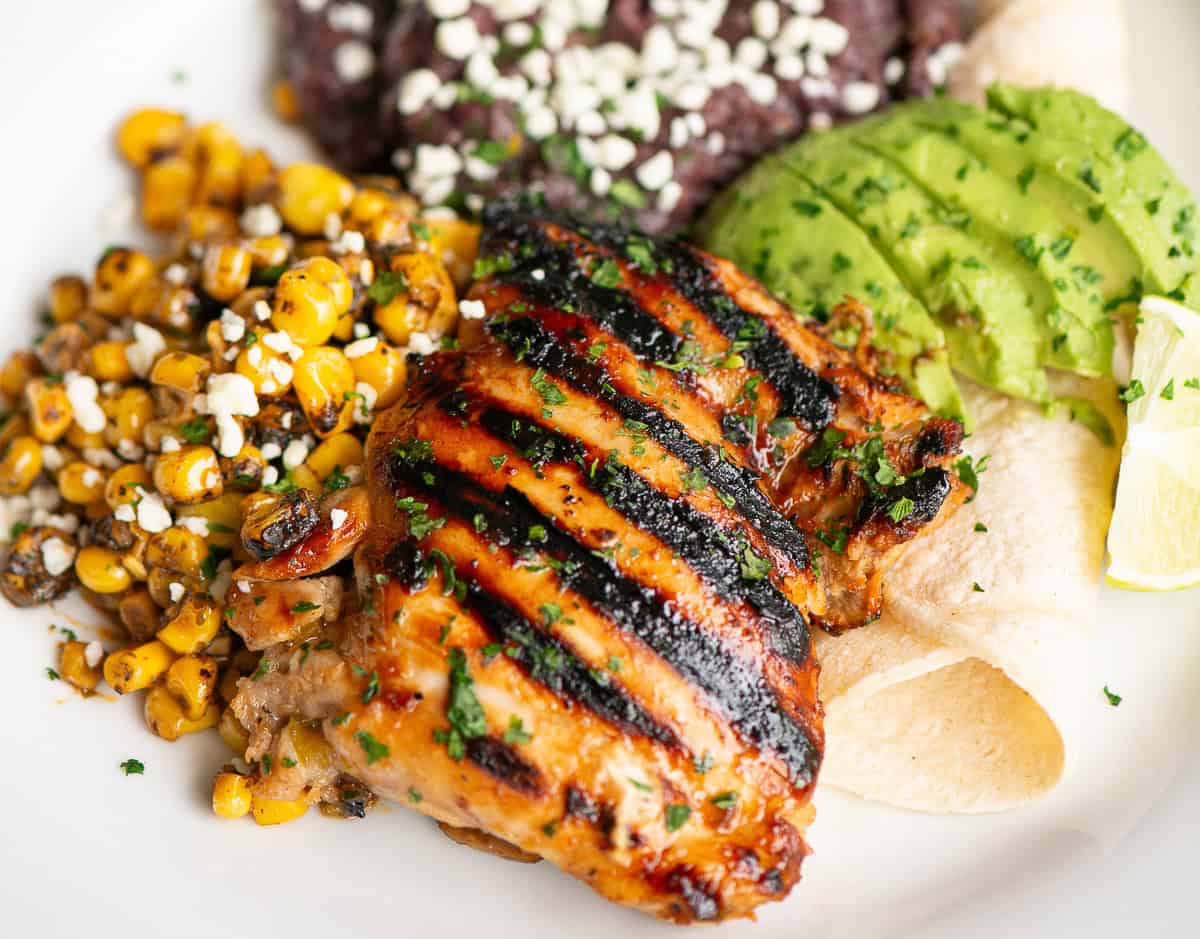 honey lime jalapeno marinated grilled chicken thigh on plate with corn, black beans, corn tortilla, and sliced avocado.