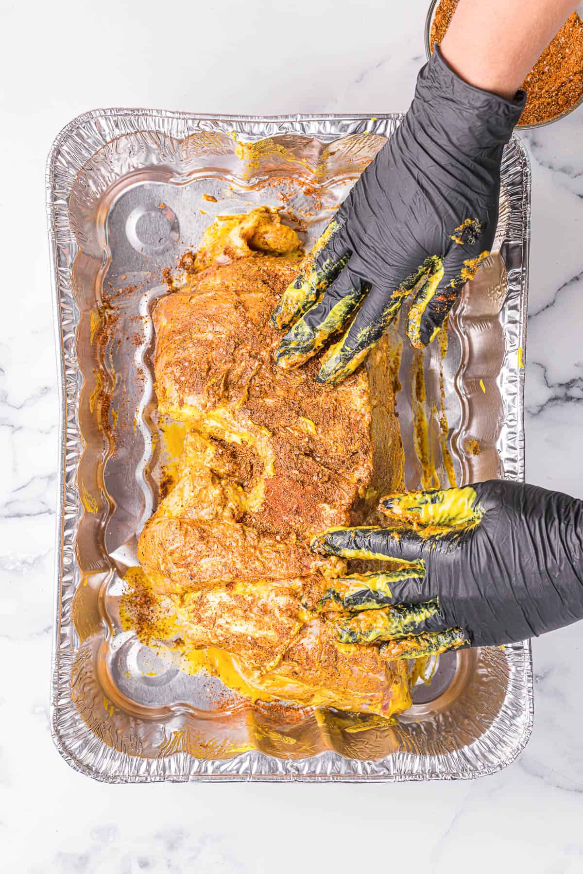 coating a pork butt with yellow mustard and dry rub seasonings with hands wearing black gloves.