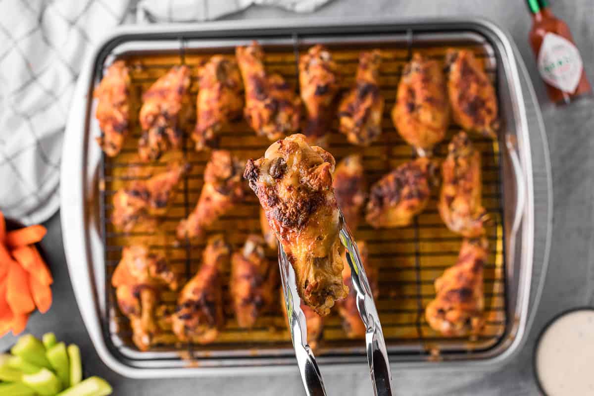 lifting up a baked ranch chicken wing with tongs over a baking sheet full of wings.