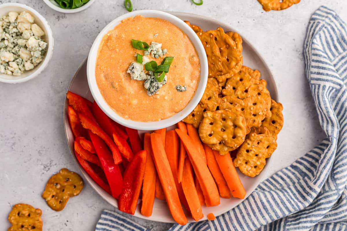 Warm buffalo chicken dip in bowl on plate with pretzel thins, carrot sticks, and sliced red bell pepper.