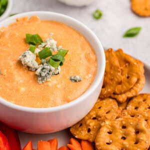 Warm buffalo chicken dip in bowl on plate with pretzel thins, carrot sticks, and sliced red bell pepper.