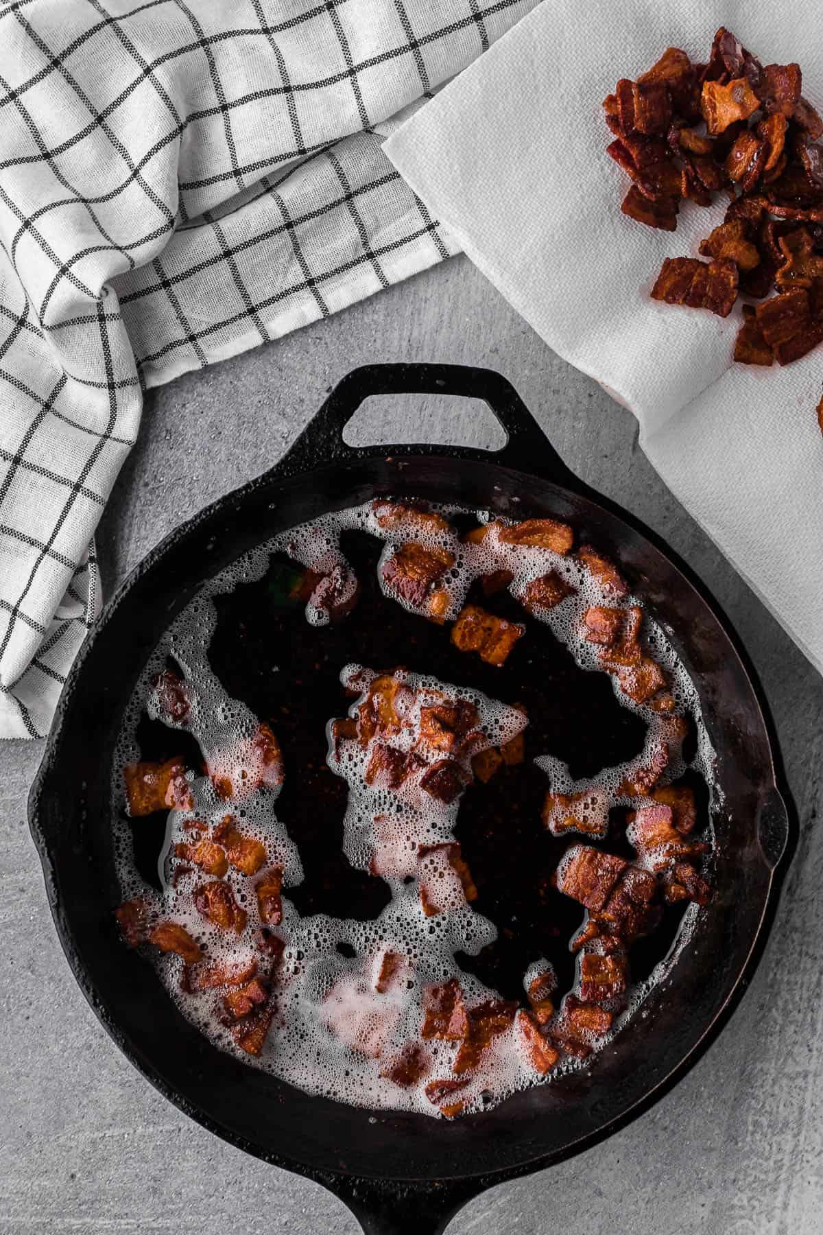 cooking bacon pieces in a cast iron skillet.