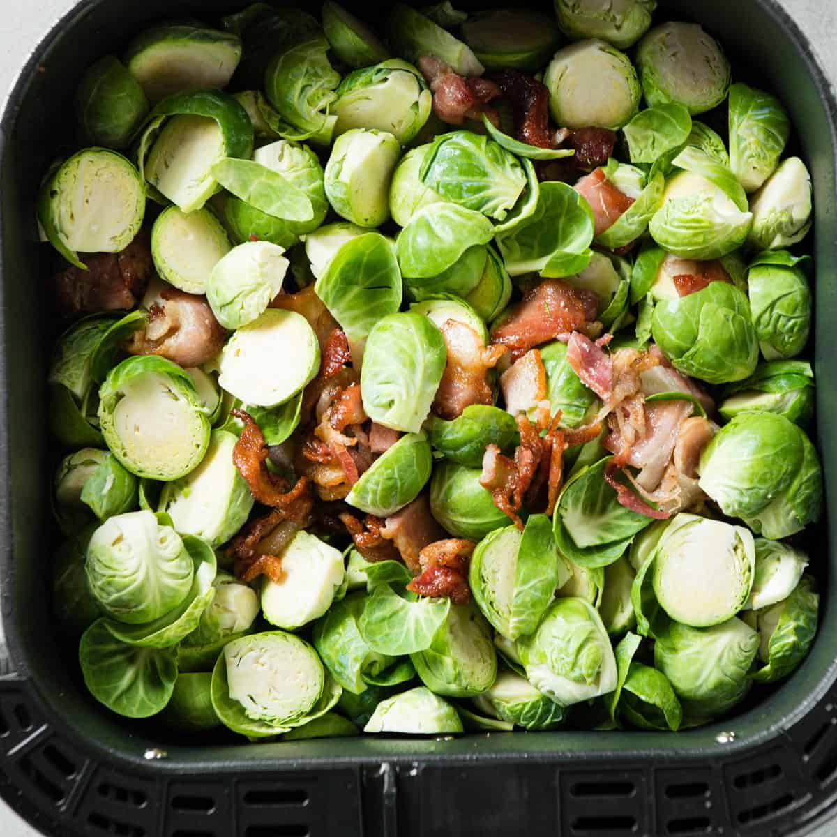 Halved Brussels sprouts and chopped bacon spread in an air fryer basket