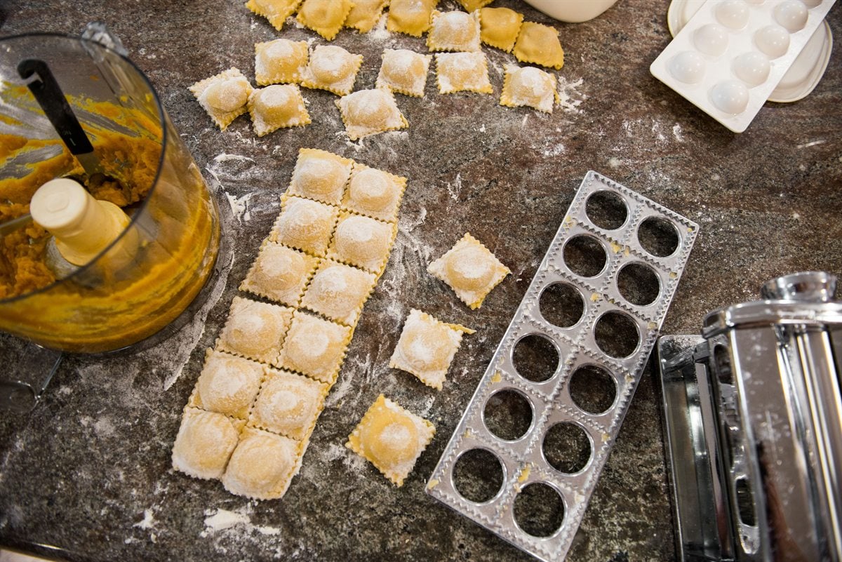 homemade ravioli on a floured counter with a ravioli press, a pasta roller and a bowl of butternut squash filling