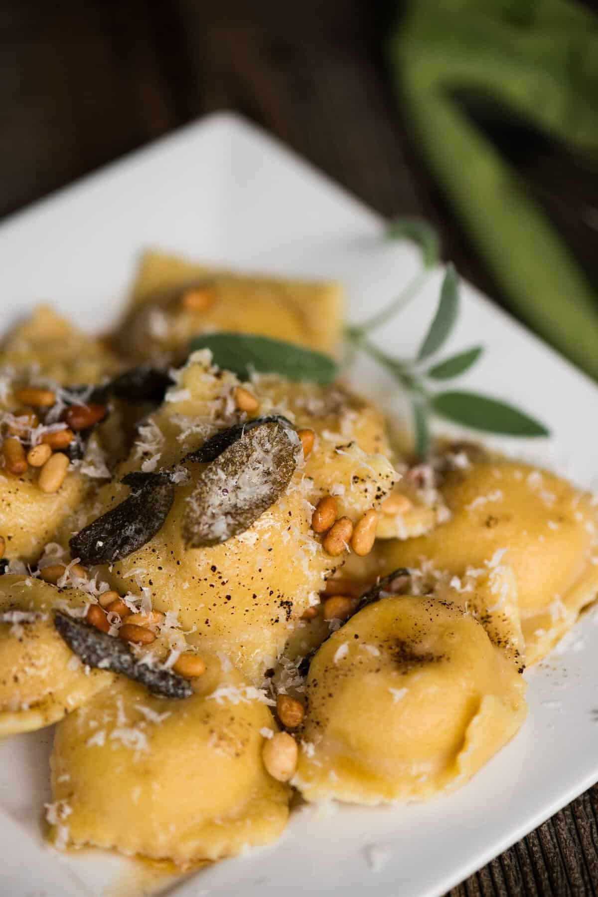 homemade butternut squash ravioli with brown butter, crispy sage leaves, toasted pine nuts and parmesan on a white plate