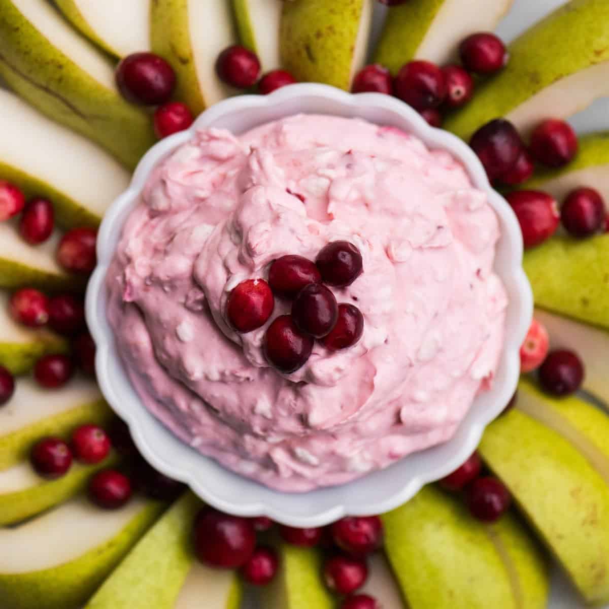 Overhead view of fluffy pink cranberry fruit dip in a scalloped white bowl, ringed with pear slices and fresh cranberries.