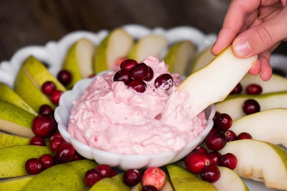 A hand dipping a pear slice into a bowl of creamy pink cranberry fruit dip.