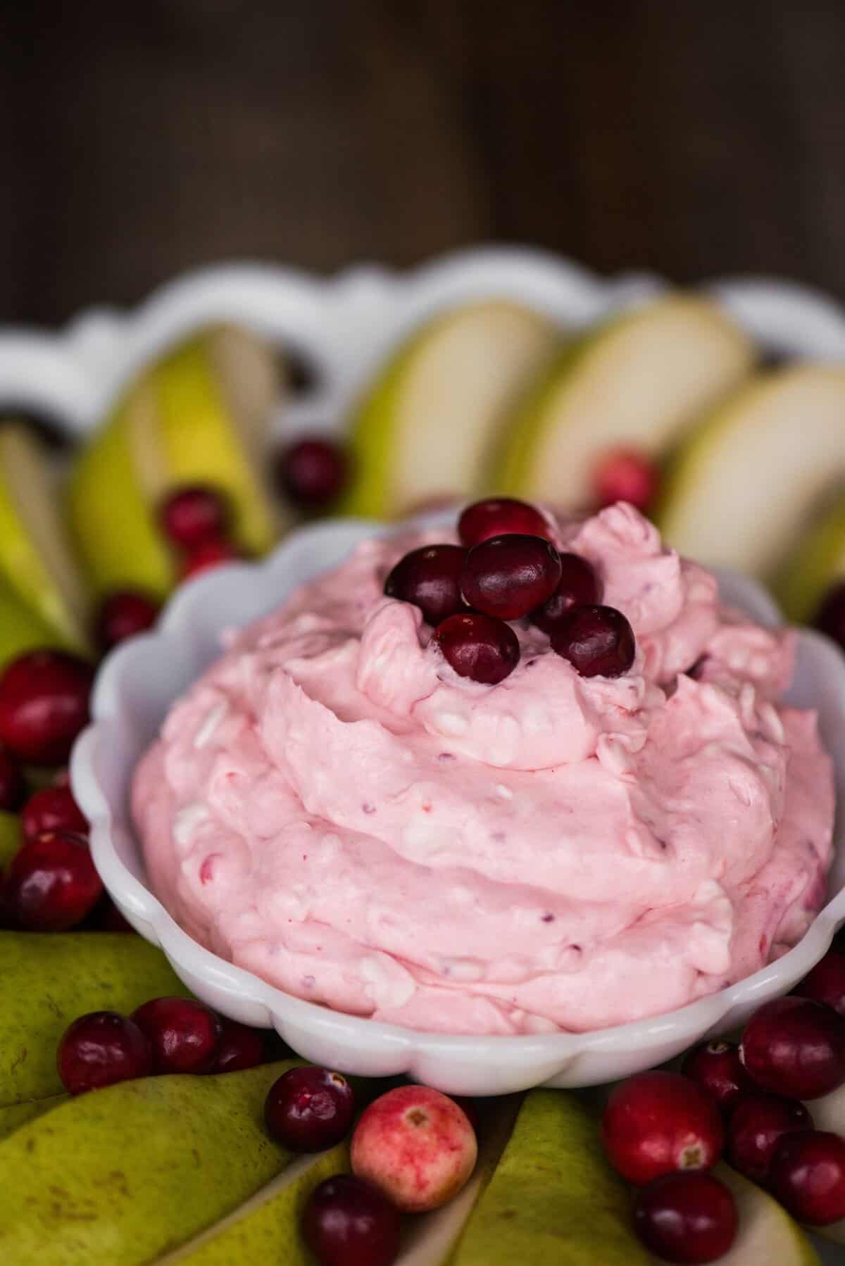 Pink cranberry fruit dip piled high in a white scalloped bowl, topped with fresh cranberries and surrounded by sliced green pears.