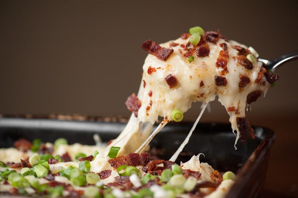 Close-up of a scoop of loaded mashed potato casserole with a long cheese pull, bacon and green onions.
