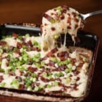 Loaded cheesy mashed potato casserole in a square baking dish with a spoonful lifted out, topped with bacon and sliced green onions.