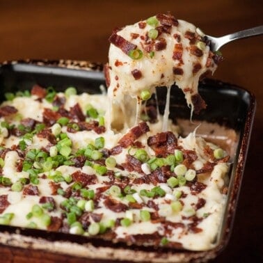 Loaded cheesy mashed potato casserole in a square baking dish with a spoonful lifted out, topped with bacon and sliced green onions.