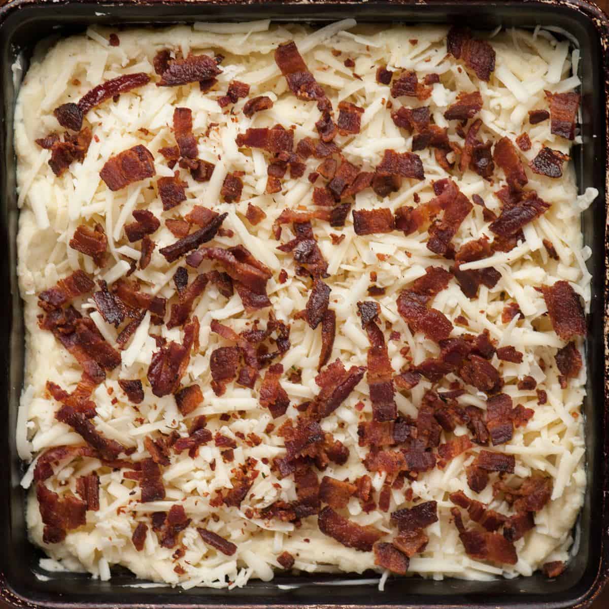 Mashed potatoes in a square baking dish topped with shredded cheddar and Gruyere and crumbled bacon, ready to bake.