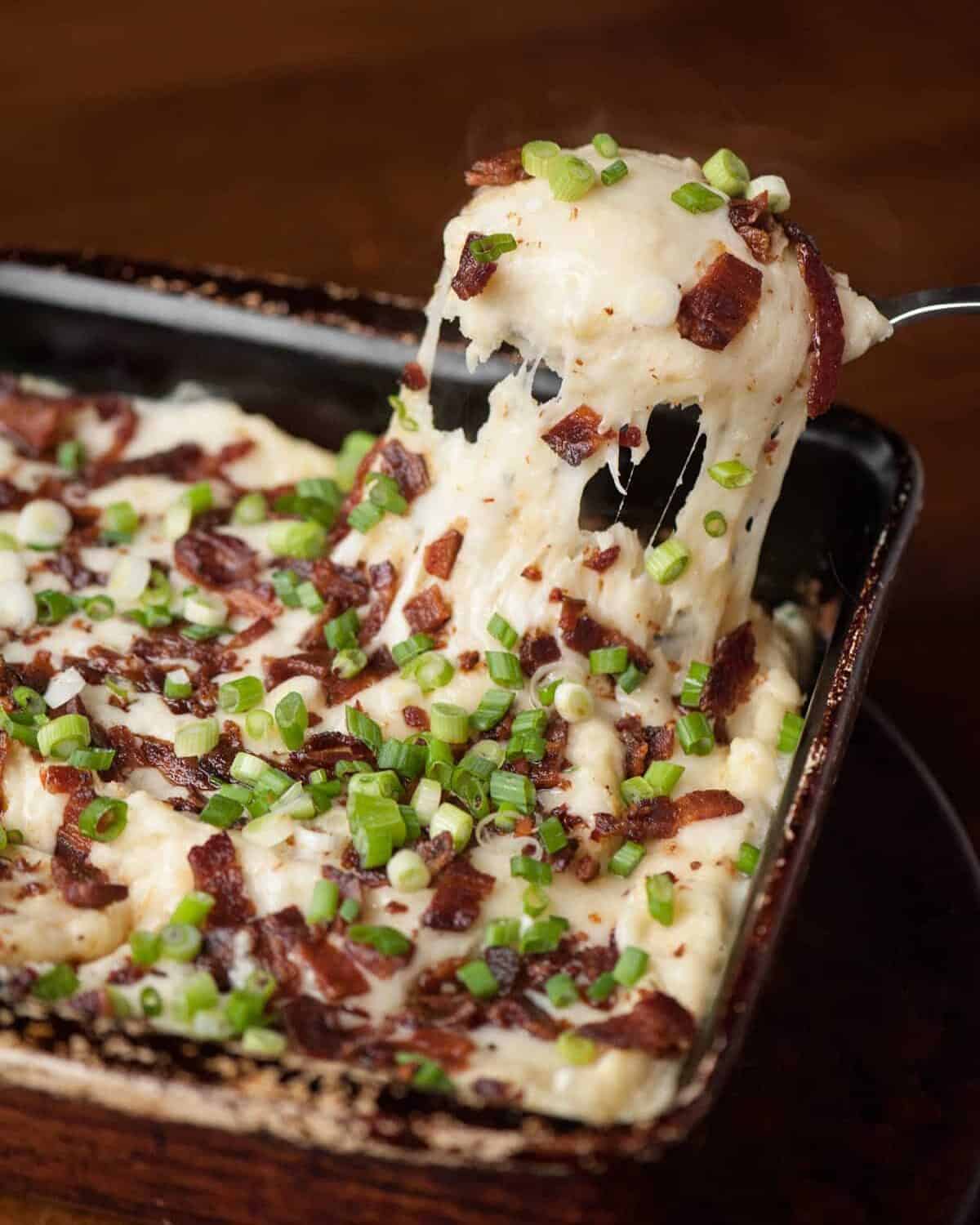 A cheesy spoonful of mashed potato casserole with crumbled bacon and green onions held above the baking dish.