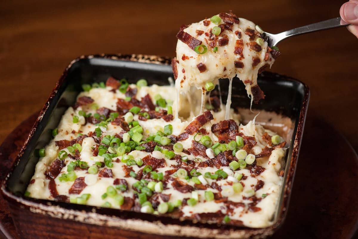 Spoonful of loaded cheesy mashed potato casserole lifted from a square baking dish, with melted cheese stretching and bacon and green onions on top.