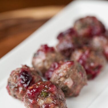 Homemade cranberry meatballs glazed in chunky cranberry chili garlic sauce, lined up on a white serving platter.