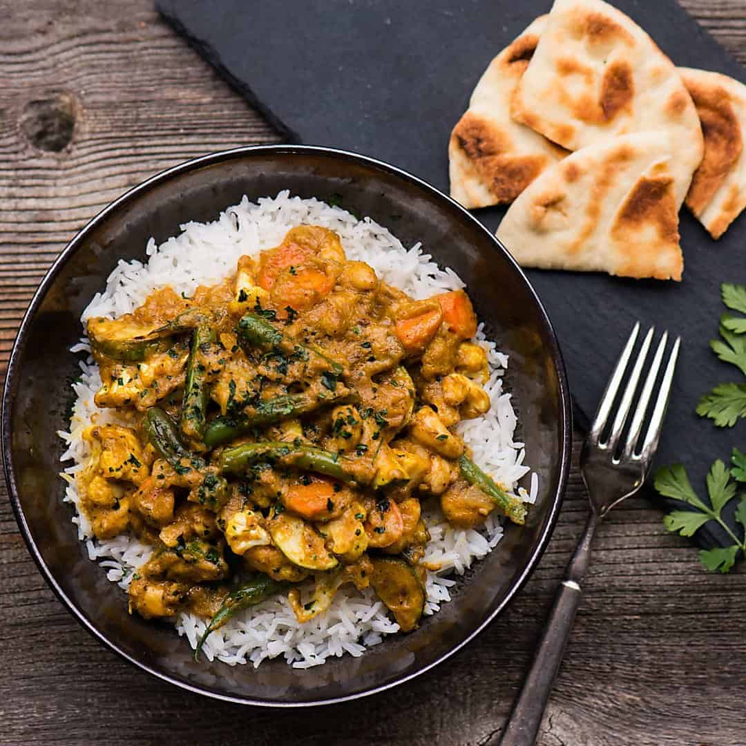 a bowl of vegetable korma with naan bread in the background