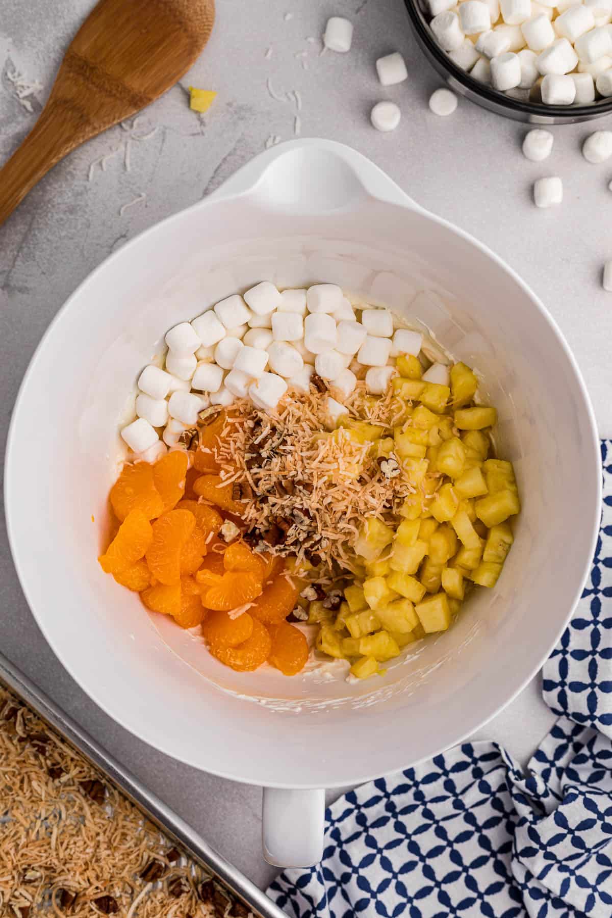 adding pineapple, oranges, marshmallows, and toasted coconut to creamy base for ambrosia salad recipe.