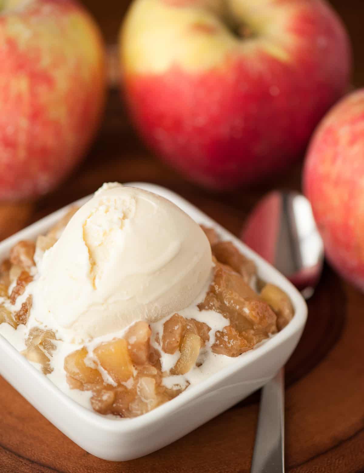 Serving of apple brown betty with vanilla ice cream on top and apples in the background.