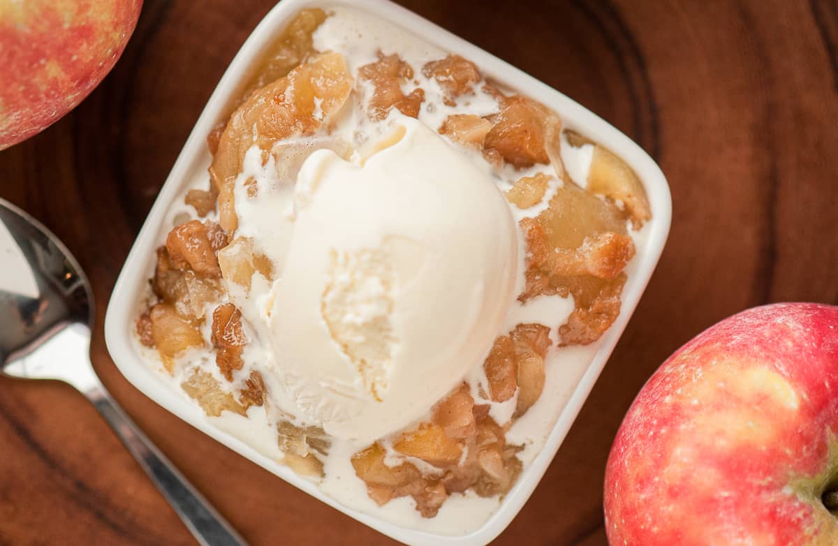 Top view of a serving of apple brown betty with vanilla ice cream on top with apples in the background.