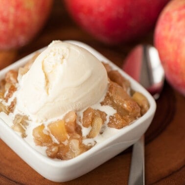 Serving of apple brown betty with vanilla ice cream on top and apples in the background.