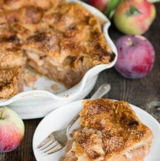 slice of apple pie on plate with whole pie and apples in background