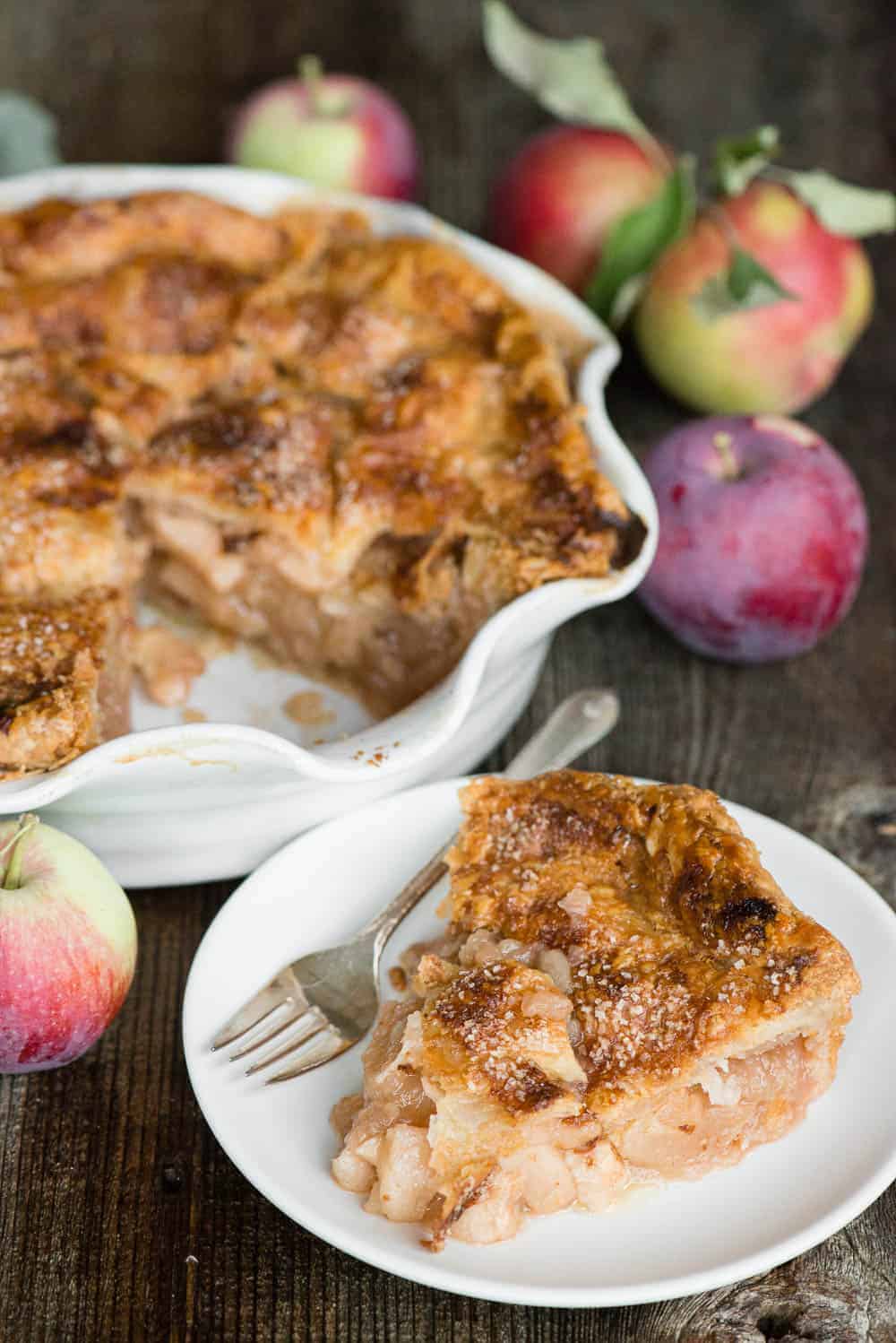 slice of apple pie on plate with whole pie and apples in background