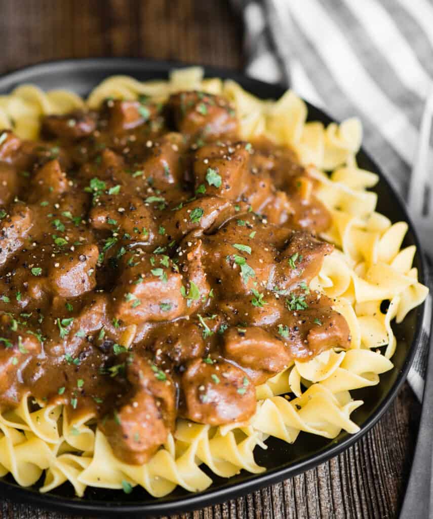 black plate with beef stew mushroom graving over cooked egg noodles.