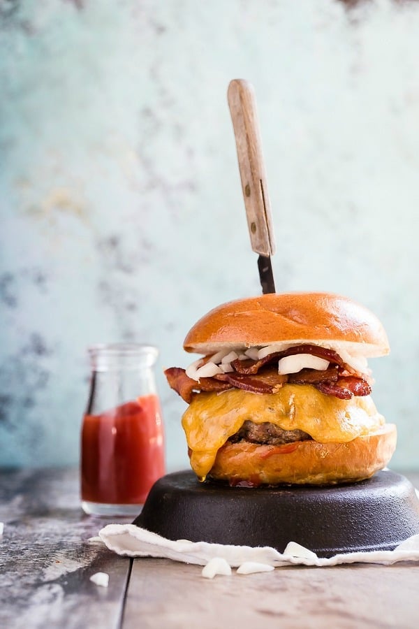 A Cheddar Bacon Cheeseburger on top of a cast iron skillet with a knife sticking out of it.