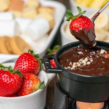 dipping strawberry into chocolate fondue with bowl of strawberries and other dippers behind.