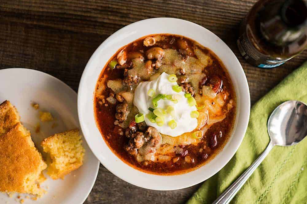 bowl of chili with cheese, sour cream, and green onions. Also, cornbread, beer, green napkin and spoon