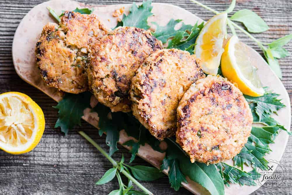 4 crispy parmesan lemon basil salmon patties on wood platter with herbs and lemon