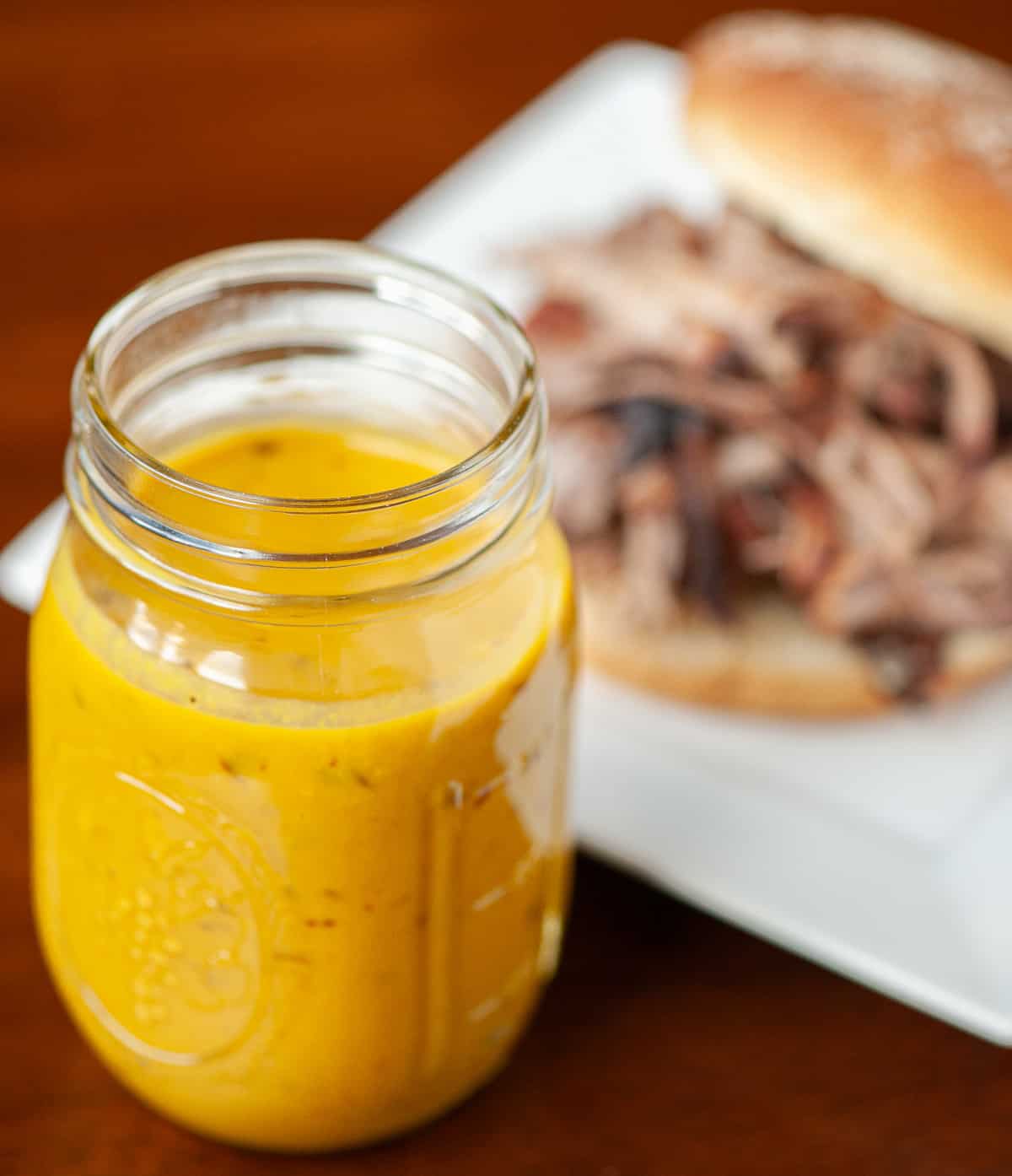 mason jar with homemade Carolina mustard bbq sauce next to pulled pork sandwich.