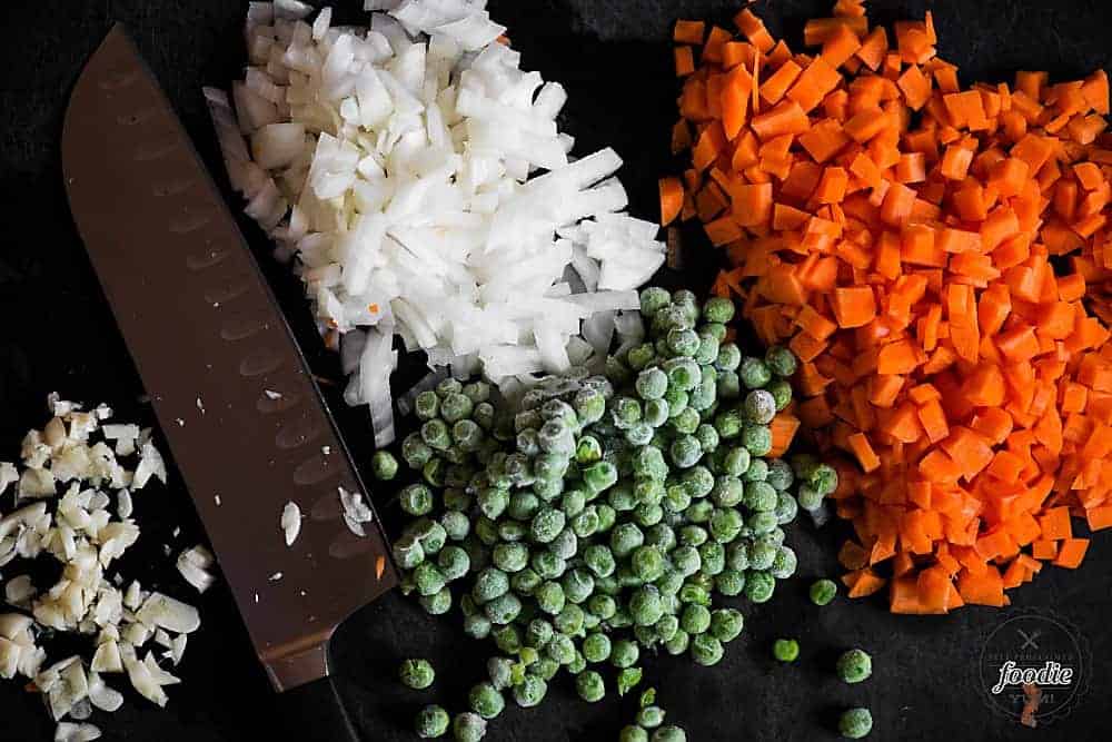 chopped onion, carrots, garlic on black cutting board with knife and frozen peas