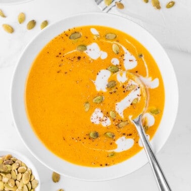 Bowl of pumpkin curry soup in white bowl garnished with heavy cream and pumpkin seeds.