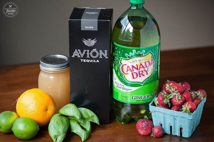ingredients to make cocktail including lemon, basil, honey, tequila, ginger ale, strawberries